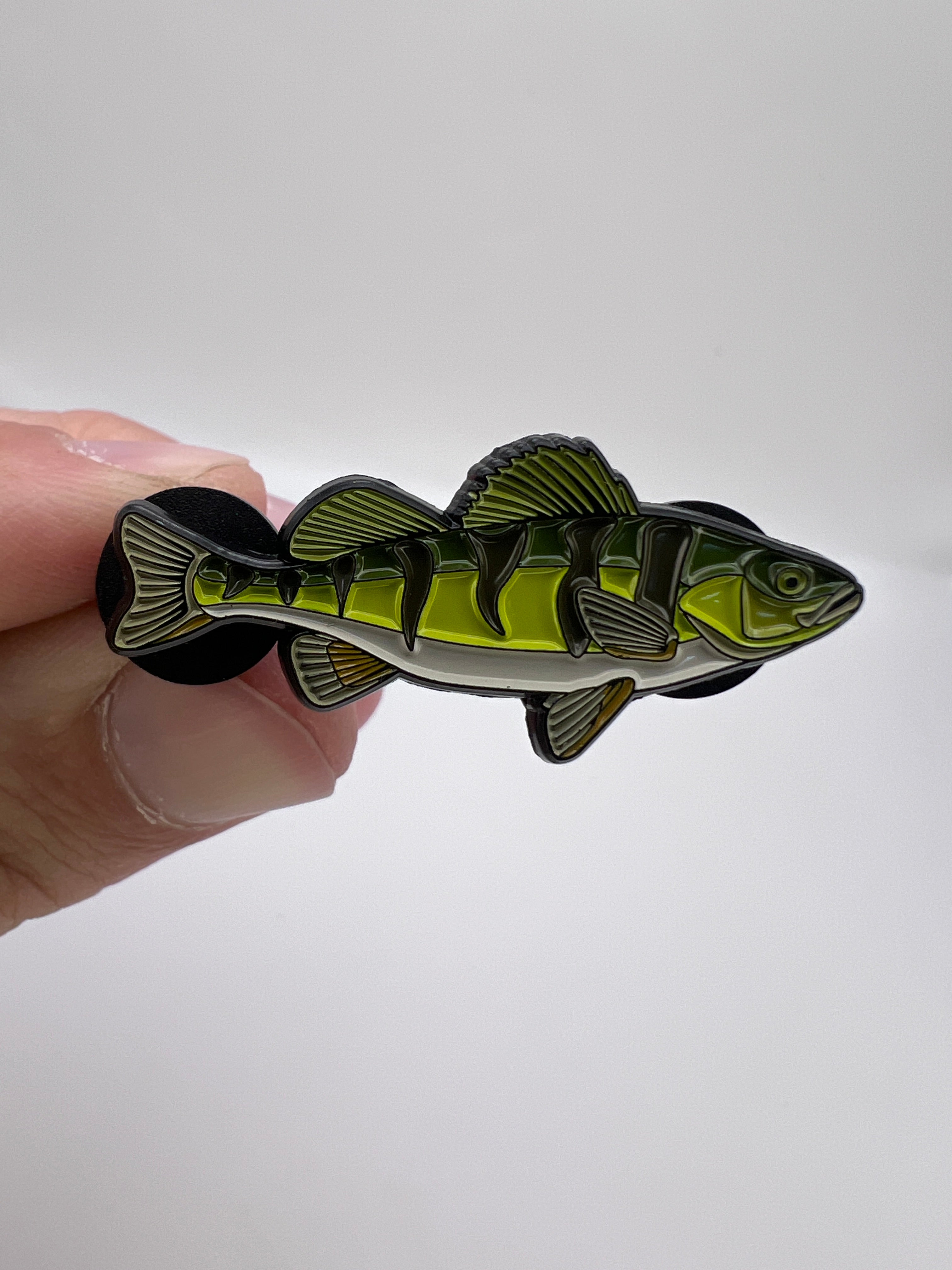 Fish-shaped green and black enamel pin held in a hand against a white background